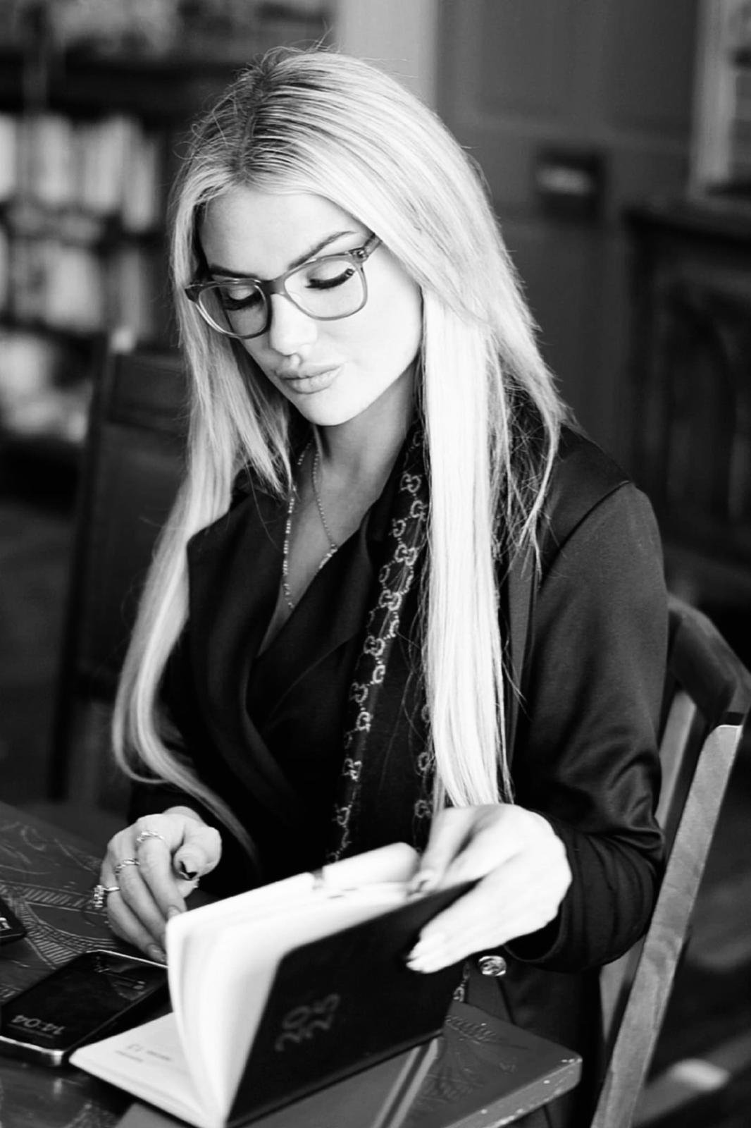 Woman with long blonde hair and glasses wearing black blazer, looking down at tablet in black and white photo